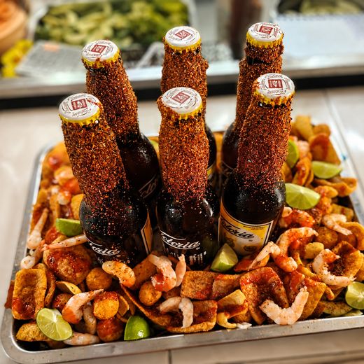 Charola con seis cervezas escarchadas de chile, rodeadas de camarón, duritos y limones